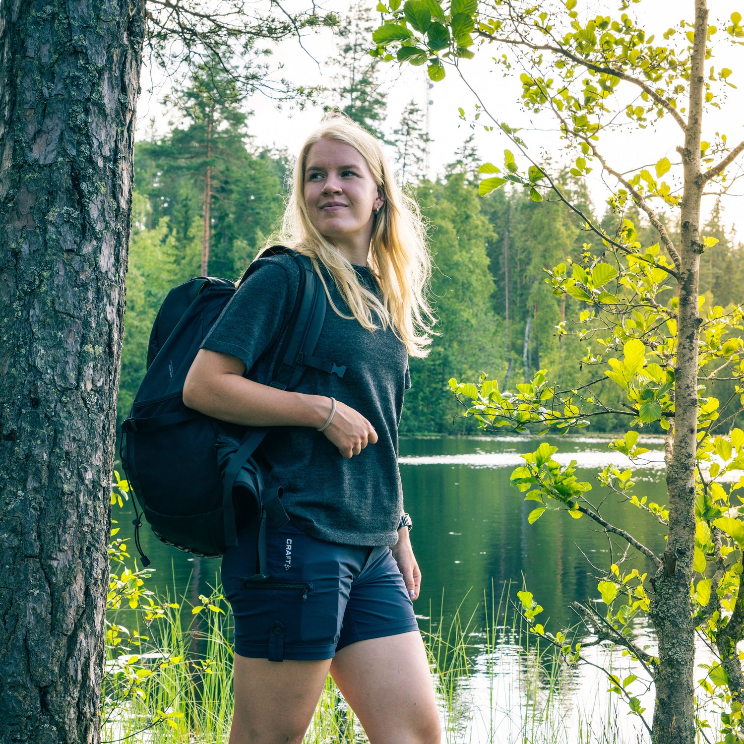 Woman with a backpack standing by a lake surrounded by trees in alpaca tee shirt